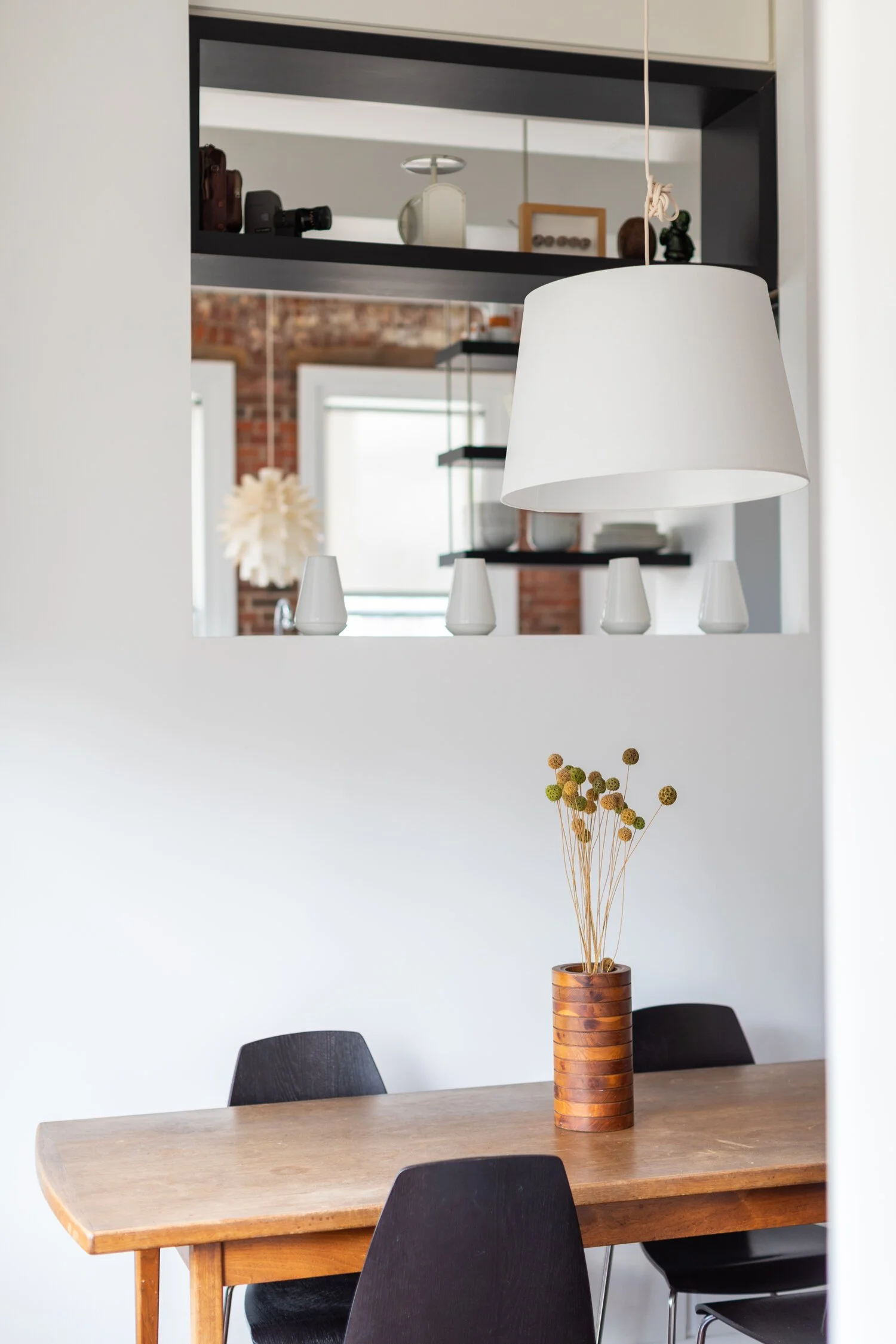 A dining room with a wooden table, black chairs, and a ceramic vase with dried flowers, seen through a cutout in a white wall. A white lampshade hangs over the table, and a mirror reflects a brick wall and windows.