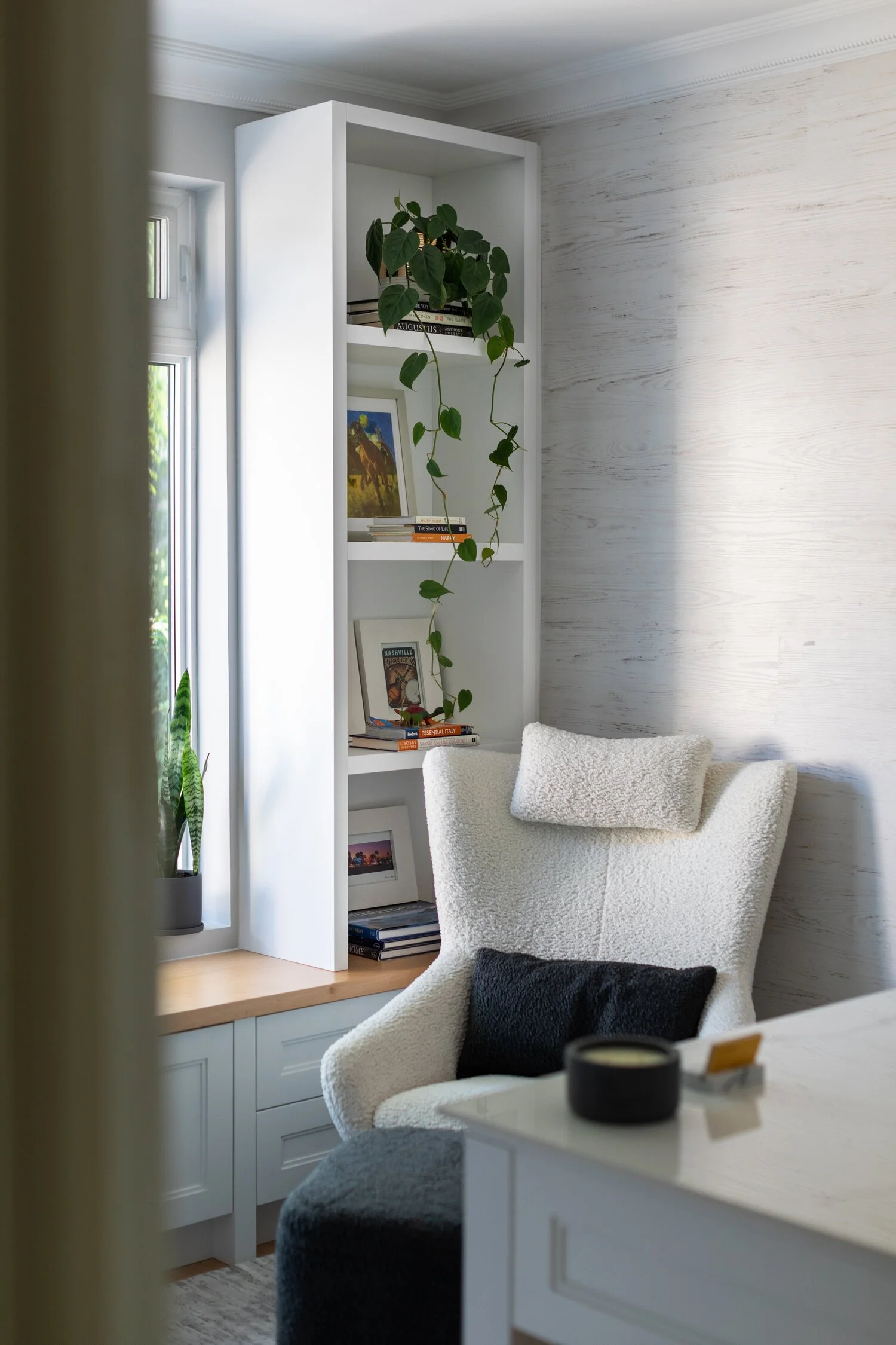A cozy corner of a room with a white, plush armchair with a pillow, near a window. There is a white bookshelf with several books and a hanging plant on top. A small potted plant sits on the window sill.