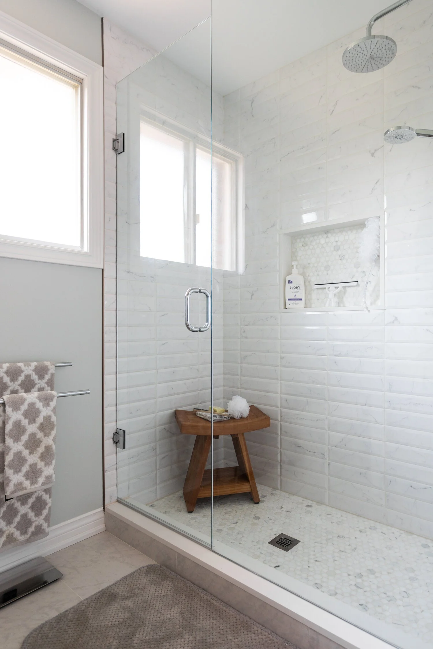 A clean, modern walk-in shower with white subway tile walls, a glass door, a rain showerhead, a small built-in niche with a bottle of soap, a wooden bench with toiletries, a window, and a beige rug outside the shower.