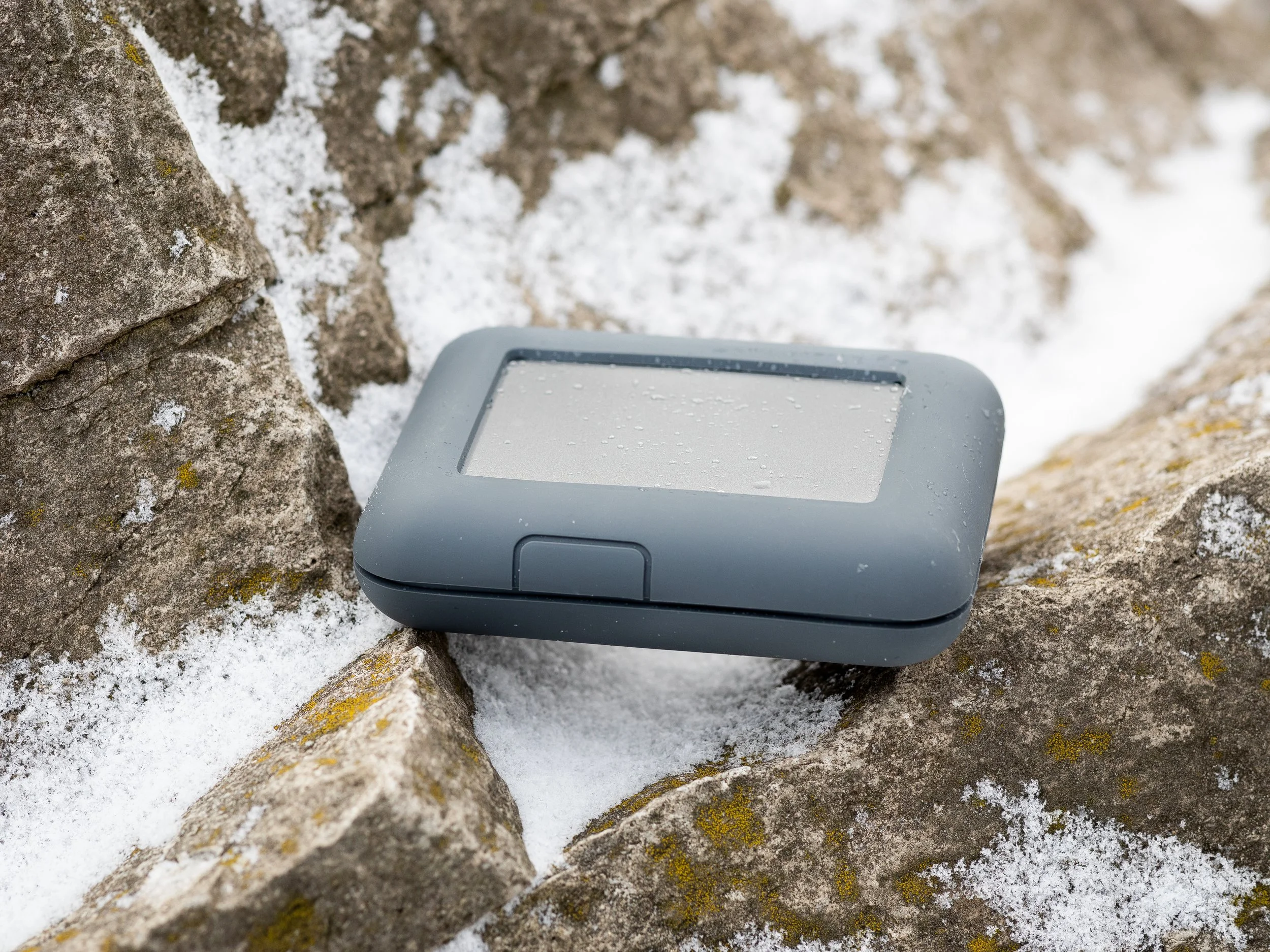 Small gray electronic device resting on snow-covered rocks.