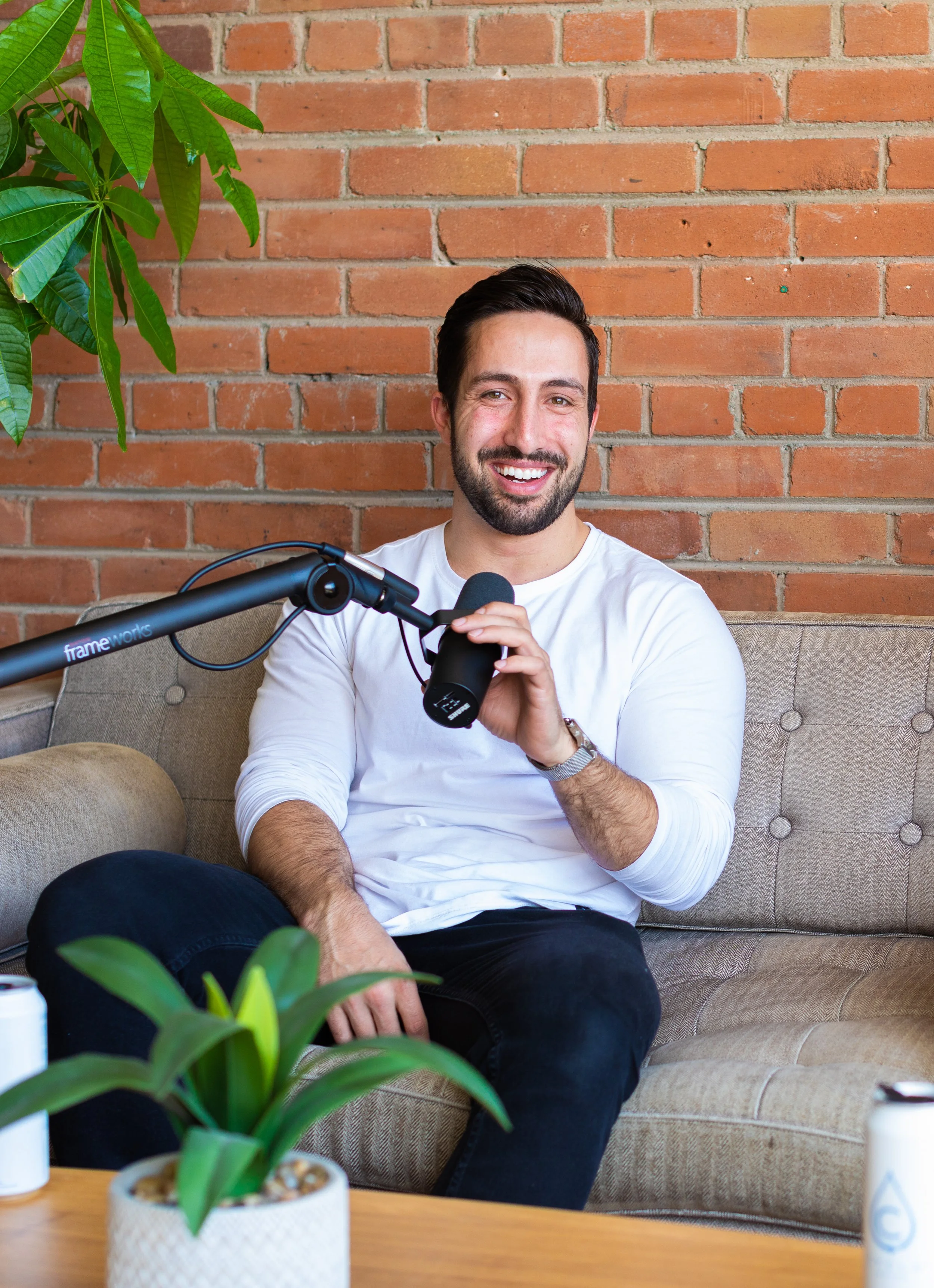 Smiling man on a beige couch speaking into a podcast microphone.