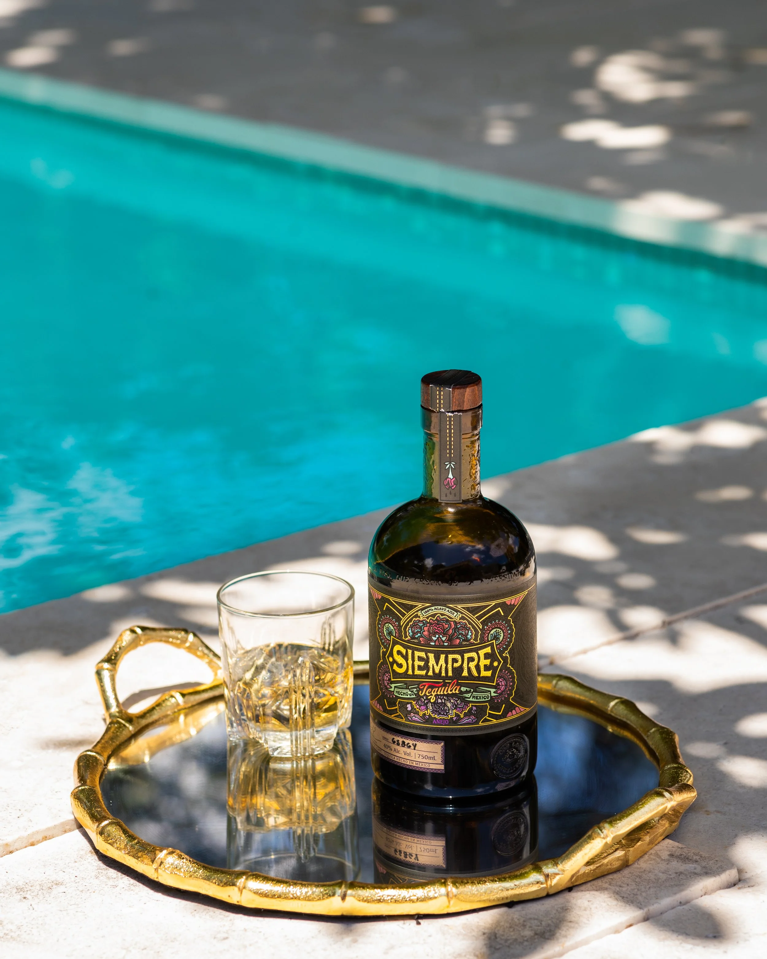 Bottle of Siempre tequila and a glass on a decorative tray beside a pool.