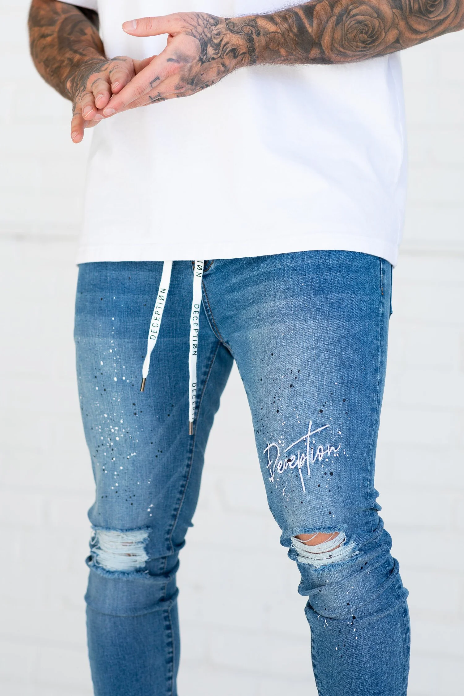 Close-up of distressed blue jeans with paint splatters and the word Deception embroidered on the thigh.