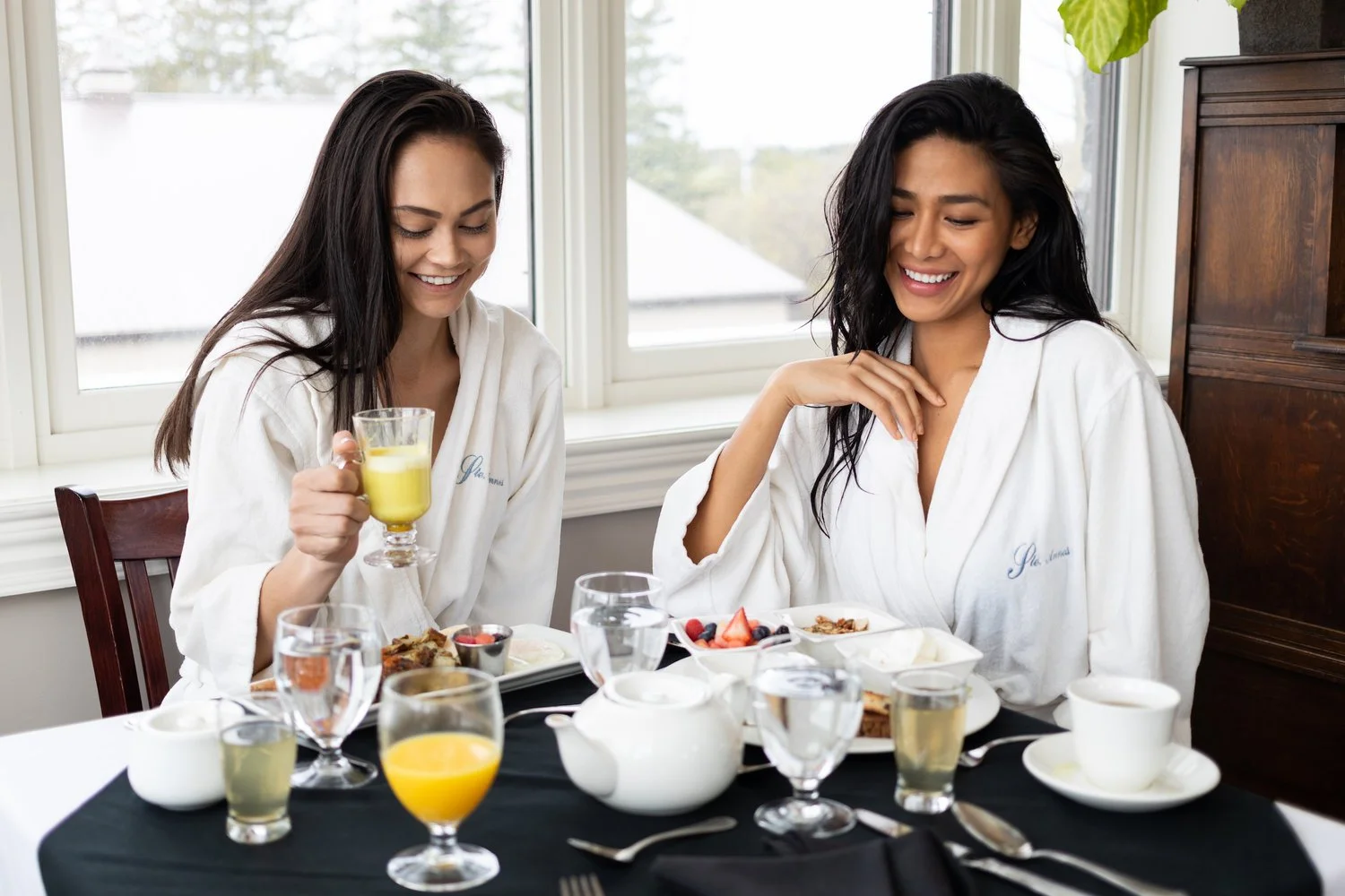 Two women in white robes enjoying breakfast at a table by large windows.