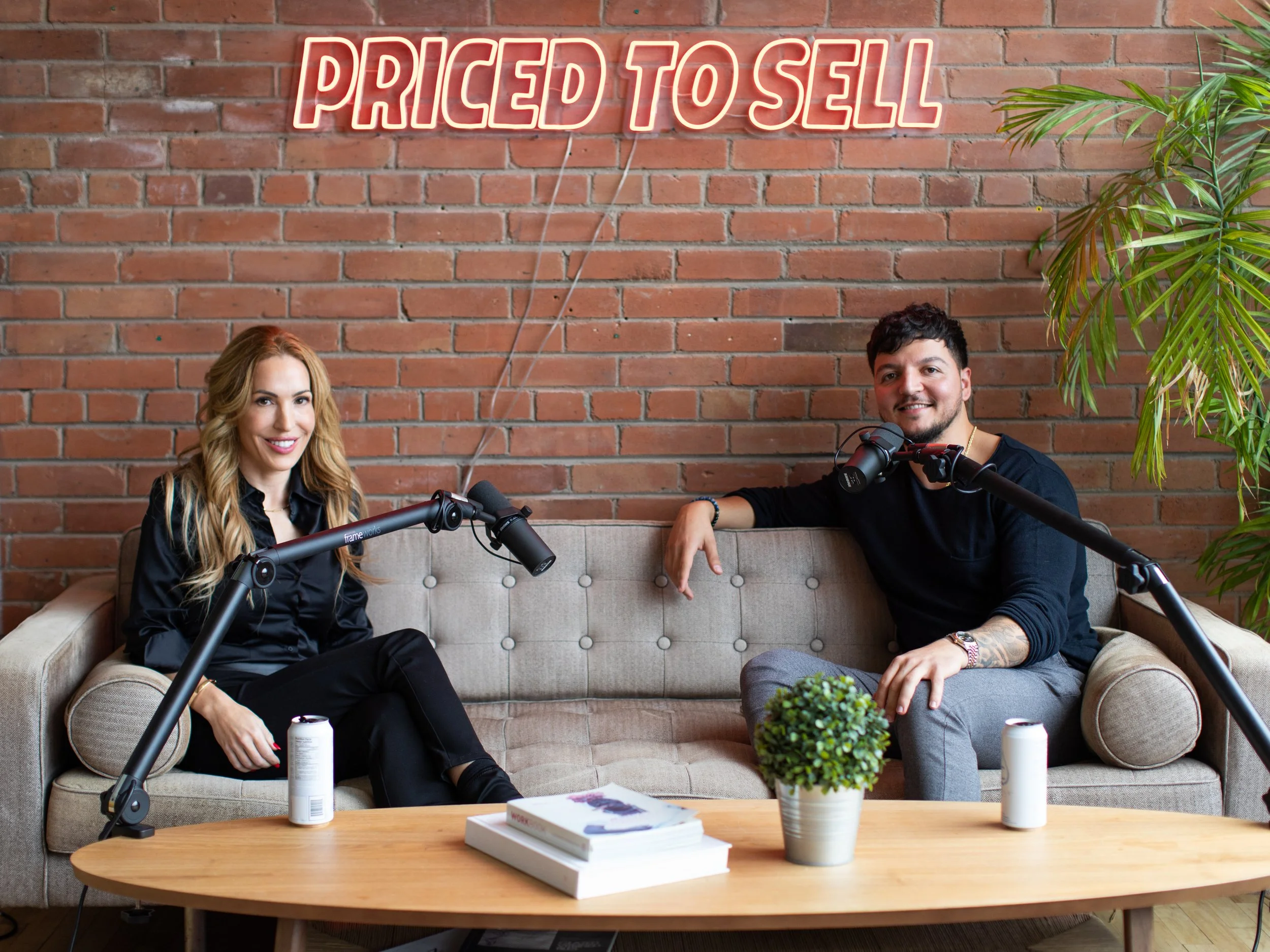 Two people recording a podcast on a beige sofa in front of a brick wall and a neon sign.