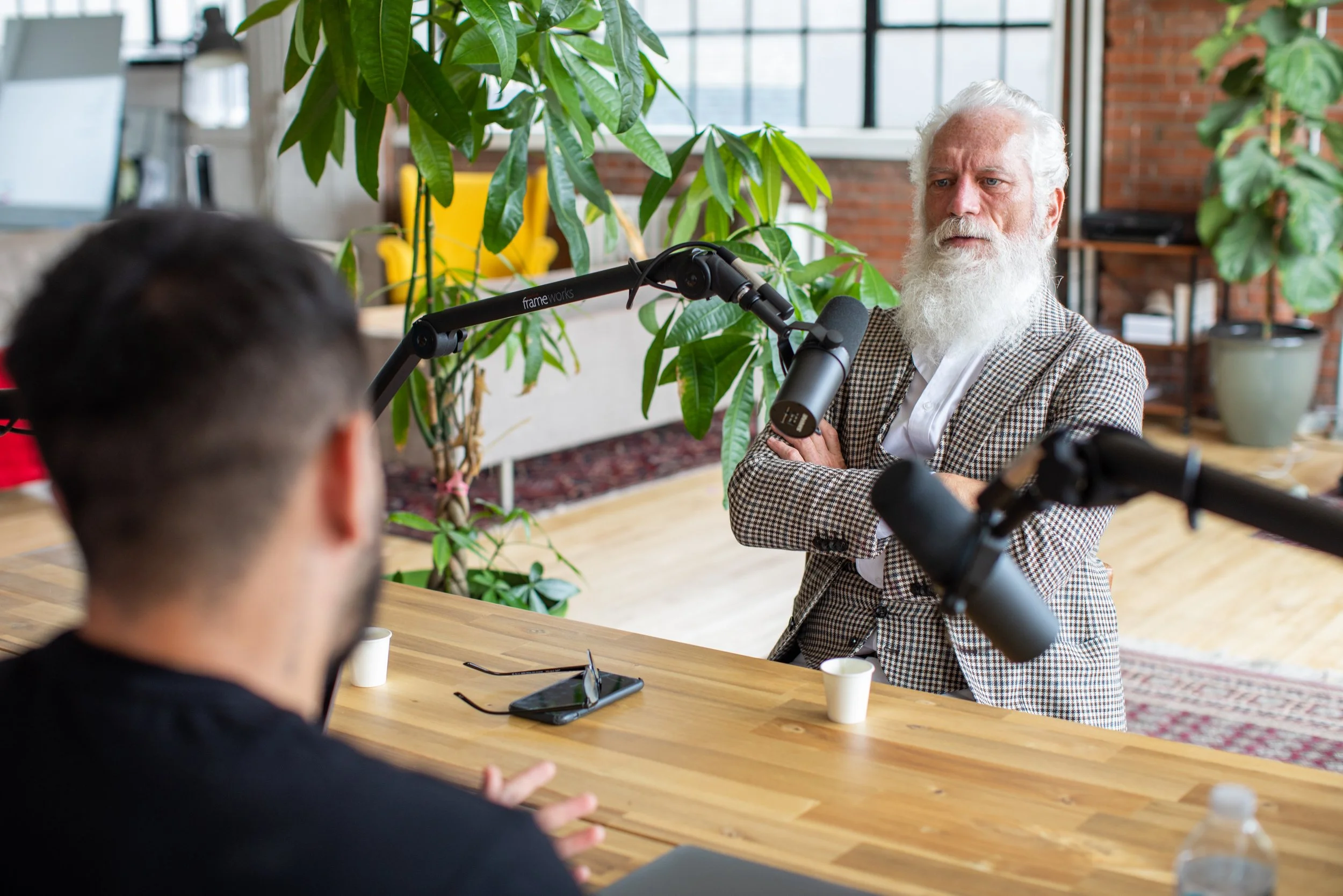 Older man in a checkered blazer speaking across a table in a bright studio setting.