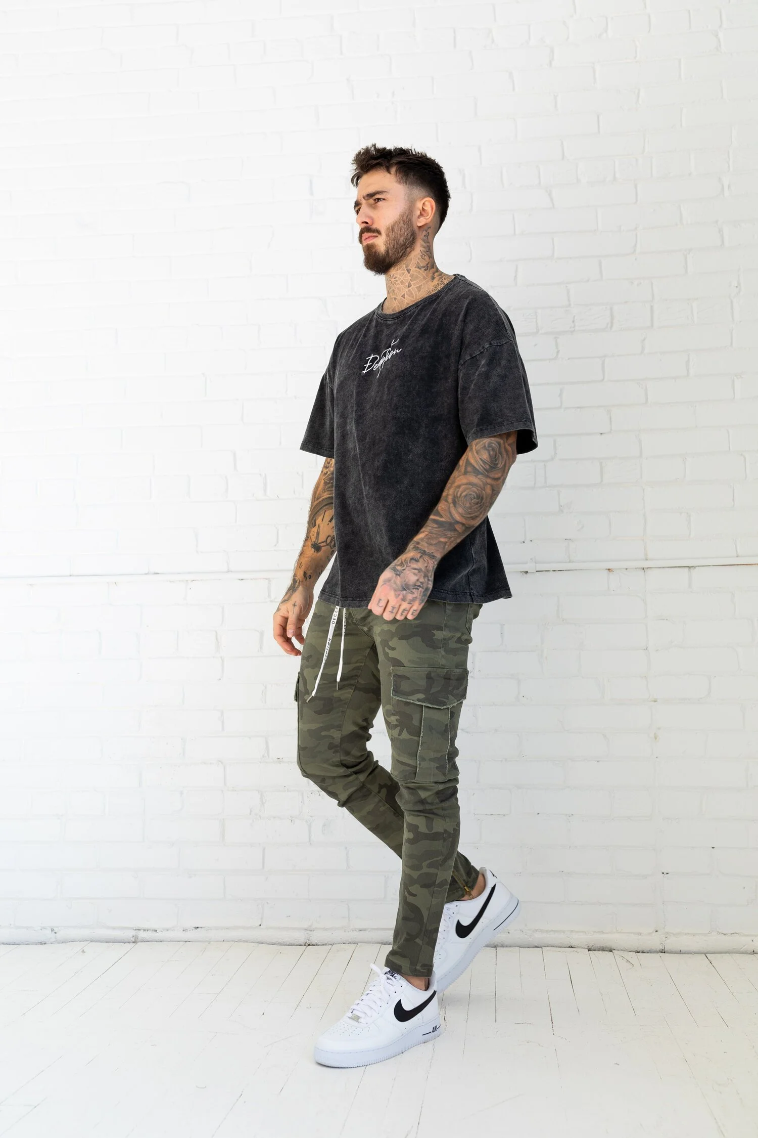 Tattooed man posing against a white brick wall in a washed black t-shirt and camo pants.
