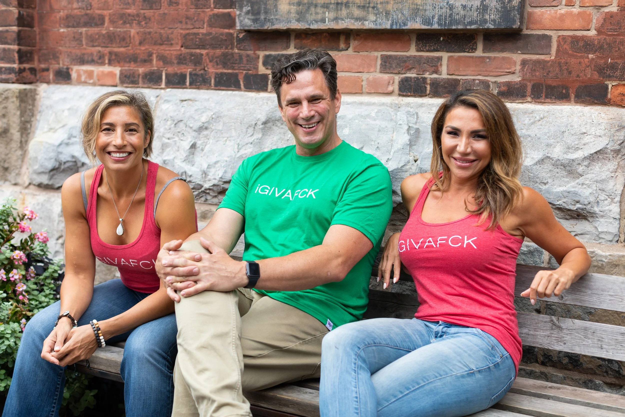 Three friends sitting on a wooden bench outdoors and smiling at the camera.