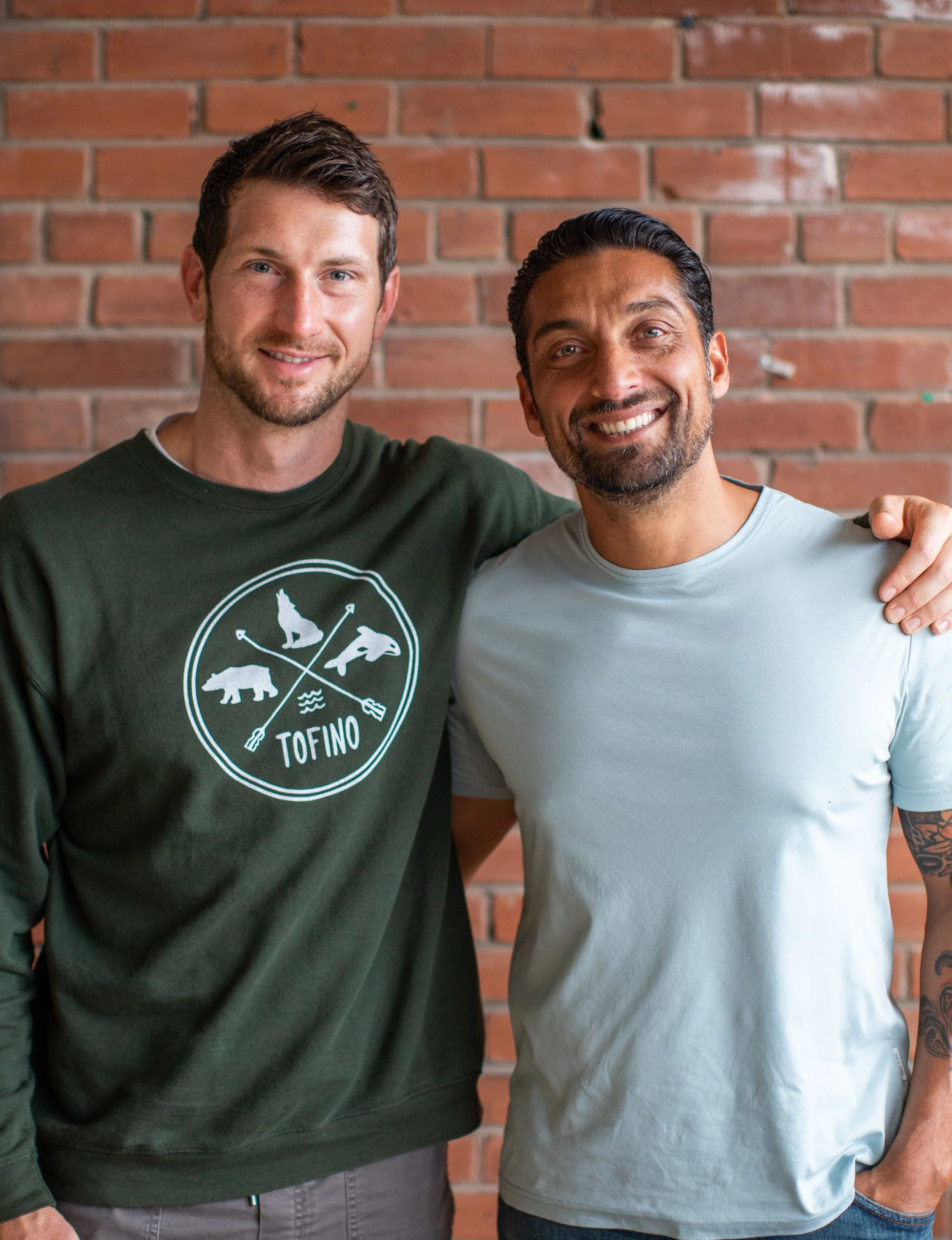 Two men smiling together in front of a brick wall.