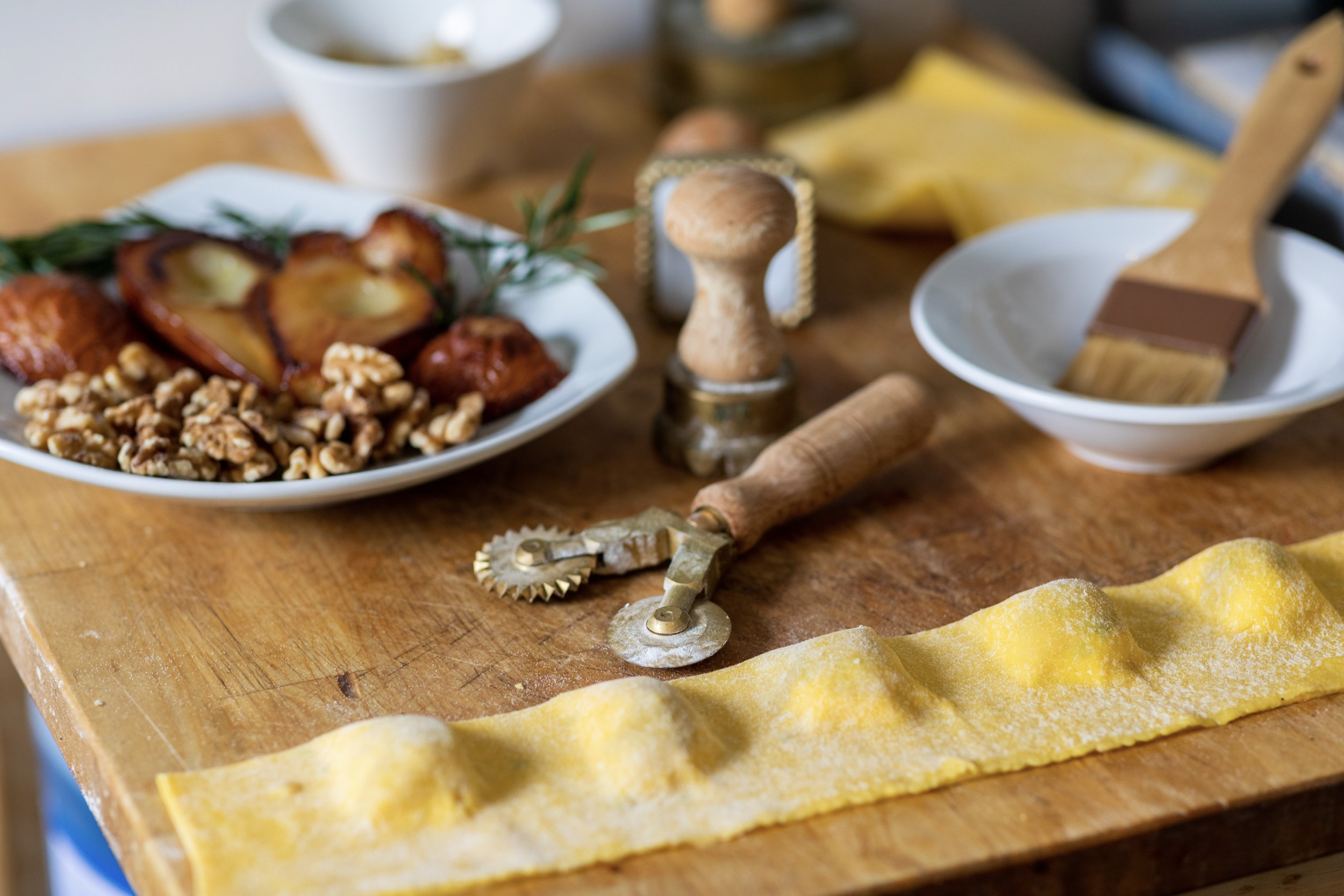 Wooden table with roasted vegetables, kitchen tools, and a rolled sheet of pasta.